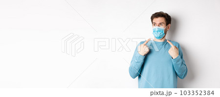 Coronavirus, health and quarantine concept. Confused young man pointing at medical mask on face and looking left, standing over white background Coronavirus, health and quarantine concept. Confused young man pointing at medical mask on face and looking left, standing over white background 103352384