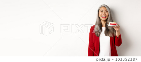 Celebration and holidays concept. Smiling asian businesswoman congratulate employees, giving plate with sweet cake and looking happy, standing over white background 103352387