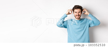 Guy put on wireless headphones to listen music or podcast, smiling at camera, standing over pink background 103352510
