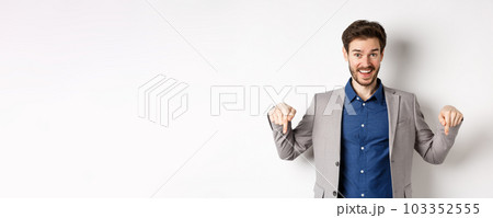 Hey check this out. Happy businessman in suit pointing fingers down and smiling, inviting to event, showing link, standing on white background 103352555