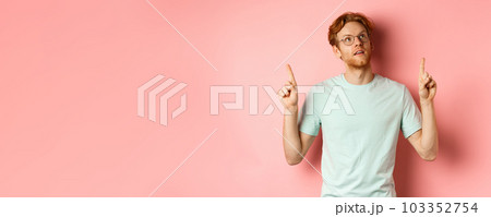 Dreamy young man with red hair and beard pointing fingers up, looking pensive at top, standing against pink background Dreamy young man with red hair and beard pointing fingers up, looking pensive at top, standing against pink background 103352754