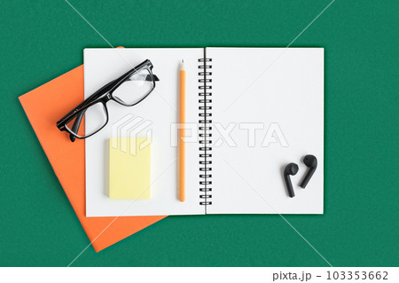 Notebook with pencil, glasses, wireless headphones. Open notebook on dark green color background, spiral notepad. Office desk. Copy Space 103353662