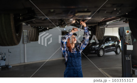 Female mechanic unscrew the nuts on the bottom of the car that is on the lift. A girl at a man's work. 103355317