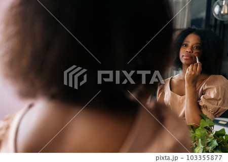 Closeup rear view from shoulder of attractive African young woman using cosmetics brush, looking to reflection on mirror. Beautiful black female putting daily makeup, applying powder at home. Closeup rear view from shoulder of attractive African young woman using cosmetics brush, looking to reflection on mirror. Beautiful black female putting daily makeup, applying powder at home. 103355877