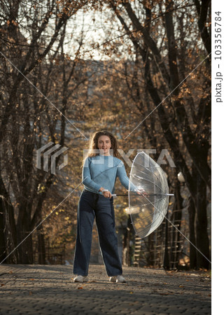 Happy beautiful woman with curly hair dance in alley of autumn park with transparent umbrella . Girl walk in park Happy beautiful woman with curly hair dance in alley of autumn park with transparent umbrella . Girl walk in park 103356784