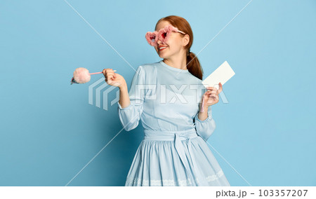 Cheerful girl, princess with red hair wearing dress and pink plush sunglasses making notes in notebook and smiling over blue studio background. Concept of education, beauty 103357207