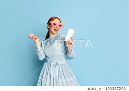 Portrait with young girl, princess with red hair wearing dress and pink plush sunglasses posing with an interested face over blue background. Concept of medieval era, beauty 103357208