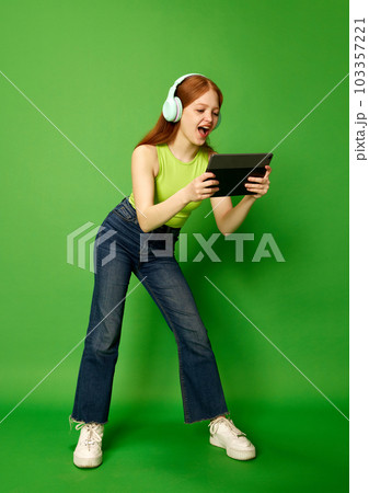 Emotive young girl, a teenager wearing headphones holding a tablet and looking at the screen, playing with interested face. Video games 103357221