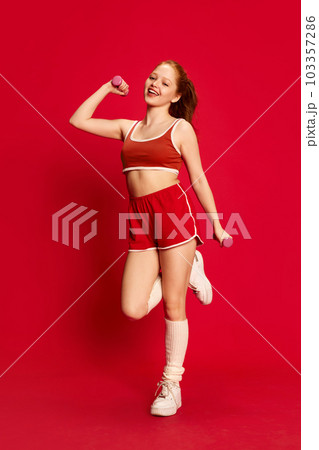 Cheerful young girl wearing sportswear doing sport exercises with pleasure over red background. Female fitness Cheerful young girl wearing sportswear doing sport exercises with pleasure over red background. Female fitness 103357286