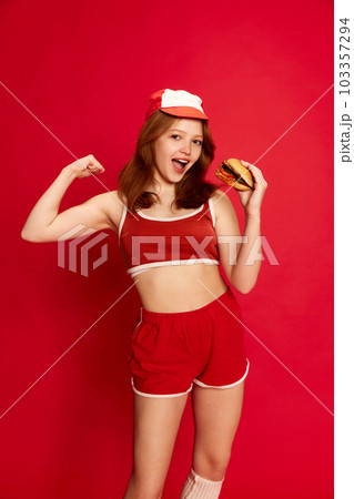 One young beautiful girl, a waitress on rollers holding big burger and demonstrating power by squeezing biceps on red background. Fast food advertising 103357294