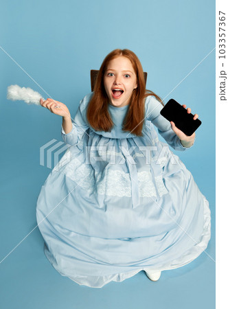 Charming princess with red hair in dress holding smartphone with surprised face over blue background. Top view. Concept of modern, technologies Charming princess with red hair in dress holding smartphone with surprised face over blue background. Top view. Concept of modern, technologies 103357367