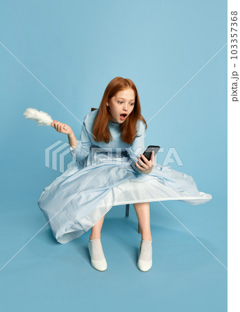 Princess, queen with red hair wearing festive dress holding smartphone with surprised face over blue background. Concept of modern, technologies, gadgets Princess, queen with red hair wearing festive dress holding smartphone with surprised face over blue background. Concept of modern, technologies, gadgets 103357368