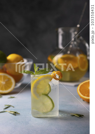 Refreshing cold summer drink with orange, lemon, lime, mint and rosemary. A misted glass with ice and citrus slices, in the background a jug with a lemonade. Close-up, selective focus. 103358431