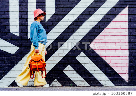 Hipster fashion young woman in bright clothes, sun glasses, bucket hat with backpack walking on the background of the painted brick wall. Urban city street fashion. Selective focus. Copy space. Hipster fashion young woman in bright clothes, sun glasses, bucket hat with backpack walking on the background of the painted brick wall. Urban city street fashion. Selective focus. Copy space. 103360597