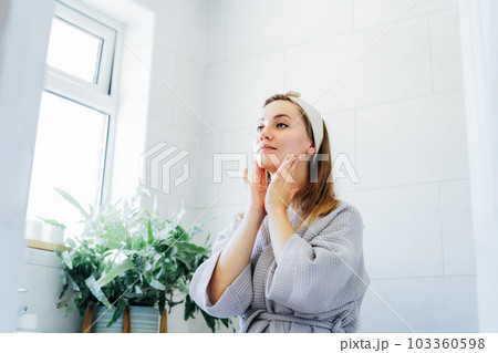Young woman in bathrobe looking in mirror and doing manual self massage of face in the bathroom. Natural procedures for skin care. Home Beauty treatment, face gymnastics for youth. Selective focus. 103360598