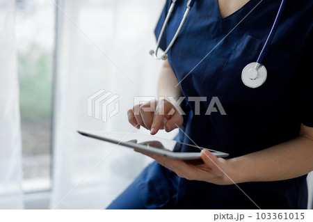 Doctor woman using tablet computer while standing near panorama window in clinic, close up. Physician or surgeon at work. Medicine concept 103361015