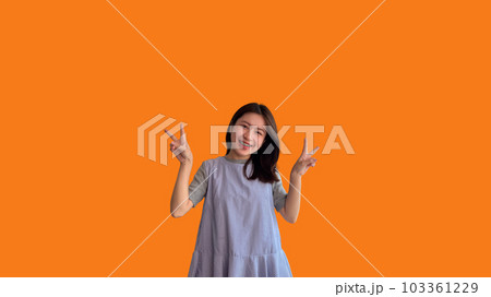 Young Asian woman making two thumbs up gesture showing joy and fun, Symbol of good friendship, Popular photo poses, Make a v-shaped finger, Orange background. 103361229