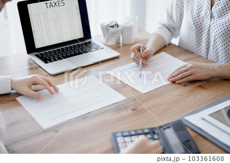 Woman accountant using a calculator and laptop computer while counting taxes with colleague at wooden desk in office. Teamwork in business audit and finance Woman accountant using a calculator and laptop computer while counting taxes with colleague at wooden desk in office. Teamwork in business audit and finance 103361608