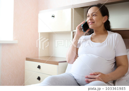 Happy curly haired multi ethnic pregnant woman, smiling looking away, gently caressing her belly while talking on mobile phone, sitting on the bed in cozy light bedroom interior. Pregnancy. Maternity Happy curly haired multi ethnic pregnant woman, smiling looking away, gently caressing her belly while talking on mobile phone, sitting on the bed in cozy light bedroom interior. Pregnancy. Maternity 103361985