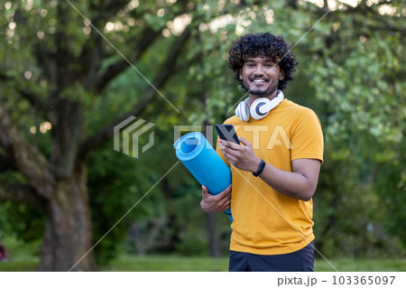 Portrait of hispanic young man standing in park wearing headphones, holding mat and phone. Engaged in physical exercises and sports. Smiling at the camera. 103365097