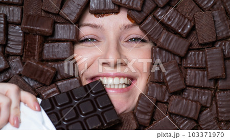 The face of a caucasian woman surrounded by chocolates. The girl eats a bar of chocolate. 103371970