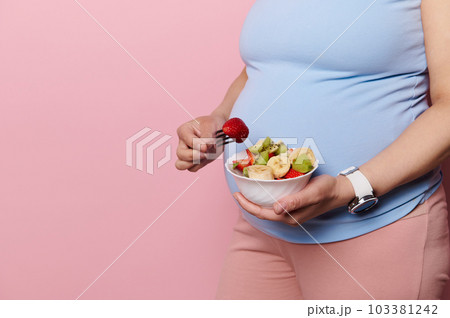 Close-up pregnant woman in blue t-shirt, holding a bowl with delicious healthy fruit salad over her big belly, isolated over pink color background. The concept of healthy nutrition in pregnancy time 103381242