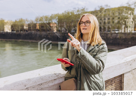 Beautiful stylish woman walking on spring day and holding mobile phone on the street background. Blonde 30s girl pointing finger. Phone Communication. Urban lifestyle concept. Check social networks 103385017