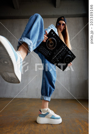 A young pretty long-haired DJ girl in a blue sweater, jeans and a black baseball cap poses with a black DJ mixing console and imitates energetic dance moves. Studio shot, gray background. 103389166