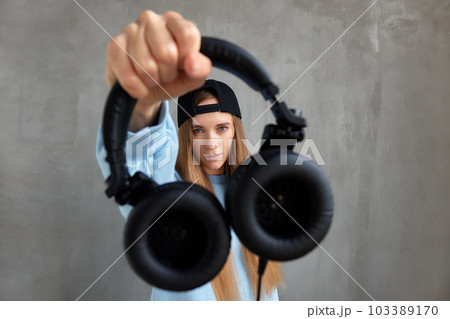 A young pretty long-haired DJ girl in a blue sweater and a funny blue hat holds black headphones in outstretched hands. Studio shot, gray background. A young pretty long-haired DJ girl in a blue sweater and a funny blue hat holds black headphones in outstretched hands. Studio shot, gray background. 103389170
