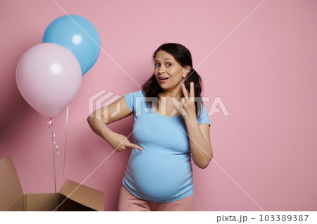 Multi ethnic middle aged glamour pretty pregnant woman, in blue t-shirt, showing at camera two fingers. Expectant mother waiting for twins, isolated on pink background with pink and blue air balloons 103389387