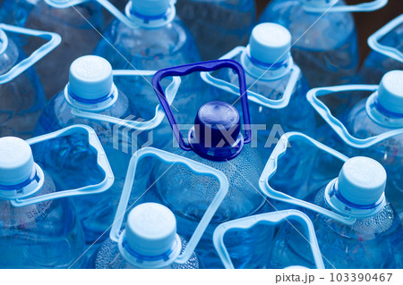 Many blue plastic bottles with blue caps and one with dark blue cap with clear mineral water in a supermarket. Pure filtered ecologically clean water in packaging. Close Up. One is different from all 103390467