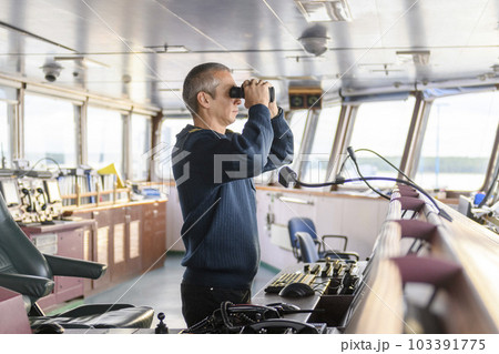 Deck officer with binoculars on navigational bridge. Seaman on board of vessel. Commercial shipping. Cargo ship. 103391775