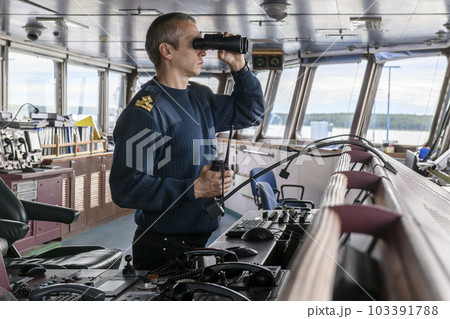Deck officer with binoculars on navigational bridge. Seaman on board of vessel. Commercial shipping. Cargo ship. 103391788