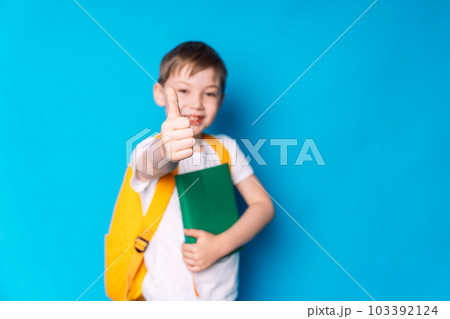 Schoolboy with a yellow backpack and a book in his hands shows a like against a blue background 103392124