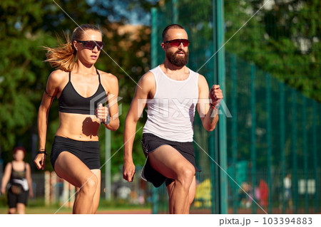 Althetes man and woman 25-35 years old, posing for the camera, smiling, running against the backdrop of a stadium with a blue sky, cardio workout, weight loss, keep yourself in good shape Althetes man and woman 25-35 years old, posing for the camera, smiling, running against the backdrop of a stadium with a blue sky, cardio workout, weight loss, keep yourself in good shape 103394883