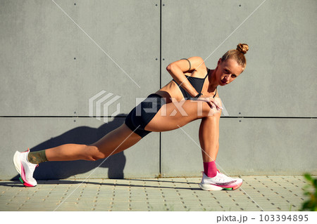 Shot of a flexible young woman warming up before running around the stadium, wearing sportswear, showing her stomach and abs, posing against a gray concrete wall 103394895