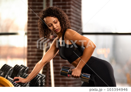 African American woman training with dumbbells in the hall in front of the mirror, against large windows, bright hall, positive view of the woman ternating the muscles of the hands African American woman training with dumbbells in the hall in front of the mirror, against large windows, bright hall, positive view of the woman ternating the muscles of the hands 103394946