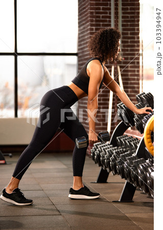 African American woman training with dumbbells in the hall in front of the mirror, against large windows, bright hall, positive view of the woman ternating the muscles of the hands 103394947