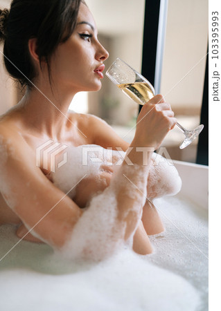 Vertical side view of alluring young woman with beautiful hairstyle and makeup drinking champagne sitting in foam bath enjoying alcoholic drink. Seductive brunette female relaxing in bathroom at home. Vertical side view of alluring young woman with beautiful hairstyle and makeup drinking champagne sitting in foam bath enjoying alcoholic drink. Seductive brunette female relaxing in bathroom at home. 103395993