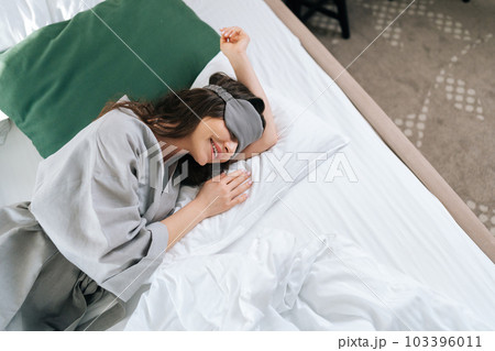 Top view of happy early bird young woman wearing sleeping mask lying in comfy bed in luxury hotel bedroom. Portrait of beautiful brunette female in pajama waking up enjoy good morning at home. Top view of happy early bird young woman wearing sleeping mask lying in comfy bed in luxury hotel bedroom. Portrait of beautiful brunette female in pajama waking up enjoy good morning at home. 103396011