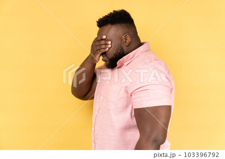 Side view of man wearing pink shirt closing eyes with hand, dont want to see that, ignoring problems, hiding from stressful situations. Indoor studio shot isolated on yellow background. Side view of man wearing pink shirt closing eyes with hand, dont want to see that, ignoring problems, hiding from stressful situations. Indoor studio shot isolated on yellow background. 103396792