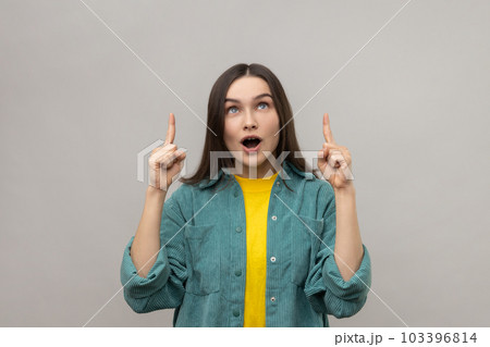 Portrait of surprised woman with open mouth pointing fingers up at empty copy space, place for advertisement, wearing casual style jacket. Indoor studio shot isolated on gray background. Portrait of surprised woman with open mouth pointing fingers up at empty copy space, place for advertisement, wearing casual style jacket. Indoor studio shot isolated on gray background. 103396814