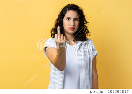 Portrait of woman with dark wavy hair showing middle finger and asking to get off looking at camera with negativity, disrespectful behaviour. Indoor studio shot isolated on yellow background. 103396817