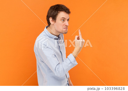 Side view portrait of angry rude man standing looking away with aggression, showing middle finger, fighting with someone, wearing light blue shirt. Indoor studio shot isolated on orange background. 103396828