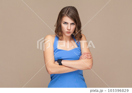 Portrait of displeased woman with wavy hair frowns face with anger, being annoyed by arguing with her boyfriend, wearing blue dress. Indoor studio shot isolated on light brown background. 103396829