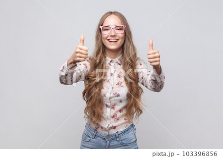 Woman in glasses with wavy blond hair looking at camera with toothy smile and showing thumbs up, approval sign, satisfied with service, good feedback. Indoor studio shot isolated on gray background. 103396856