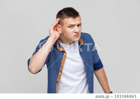 Portrait of teenager boy wearing blue shirt holding hand near ear and listening carefully, having hearing problems, deafness in communication. Indoor studio shot isolated on gray background. Portrait of teenager boy wearing blue shirt holding hand near ear and listening carefully, having hearing problems, deafness in communication. Indoor studio shot isolated on gray background. 103396863