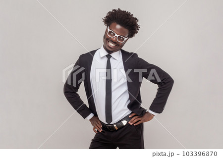 Portrait of funny positive man in glasses standing with hands on hips, looking at camera with toothy smile, wearing white shirt and tuxedo. Indoor studio shot isolated on gray background. Portrait of funny positive man in glasses standing with hands on hips, looking at camera with toothy smile, wearing white shirt and tuxedo. Indoor studio shot isolated on gray background. 103396870