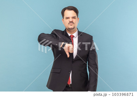 Displeased sad showing man with mustache standing showing dislike gesture, grimacing, feels dissatisfied, wearing black suit with red tie. Indoor studio shot isolated on light blue background. 103396871