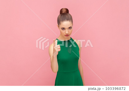 Portrait of serious strict young adult woman standing with bossy facial expression, raising fore finger, warning you about problems, wearing green dress. Indoor studio shot isolated on pink background Portrait of serious strict young adult woman standing with bossy facial expression, raising fore finger, warning you about problems, wearing green dress. Indoor studio shot isolated on pink background 103396872